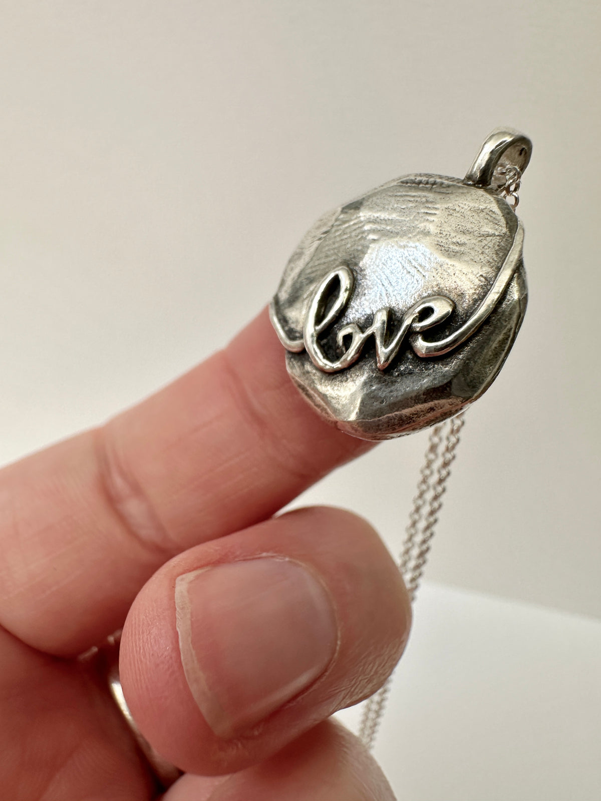 Round silver pendant with 'love' written across it, it is domed with a rough texture - being held on the tip of a finger.